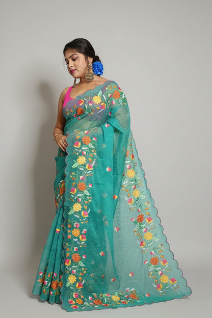 Woman wearing a teal saree with floral patterns, standing against a plain background.