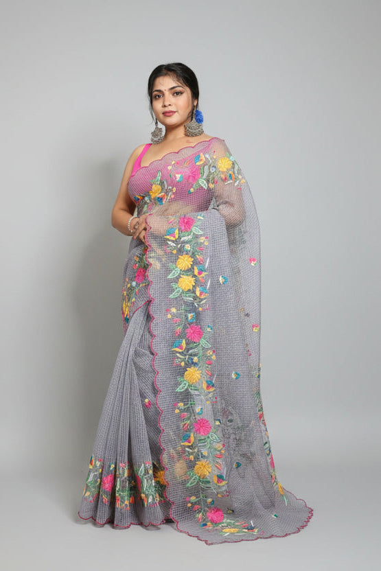 Woman in a saree with floral patterns standing next to traditional pots against a dark background.