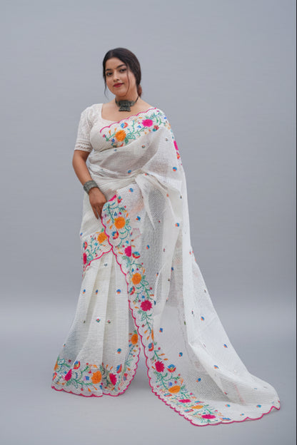 Woman wearing a white saree with colorful floral embroidery against a green outdoor background